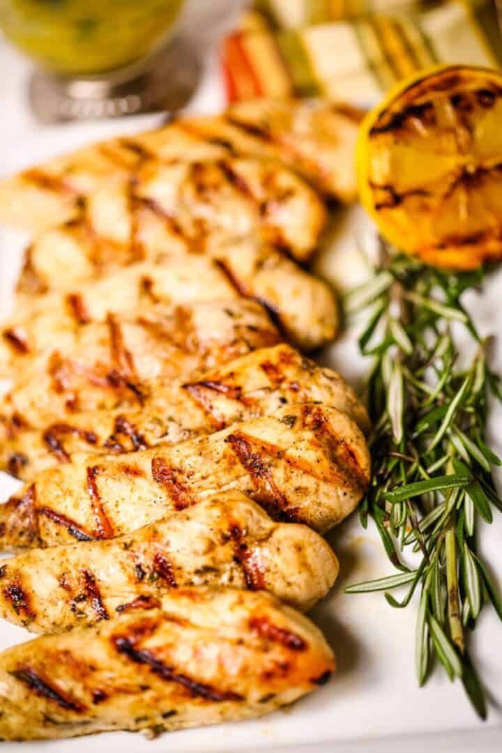 Breasts are arranged on a plate with fresh rosemary, grilled lemon, and sliced vegetables in the background.