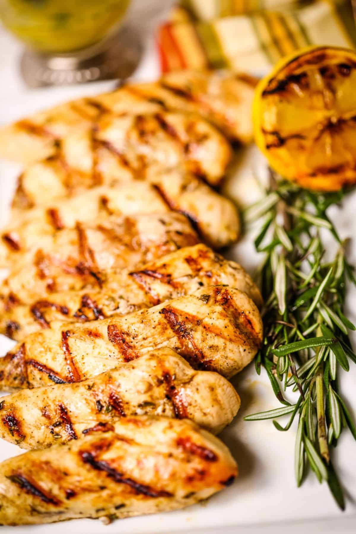 Breasts are arranged on a plate with fresh rosemary, grilled lemon, and sliced vegetables in the background.