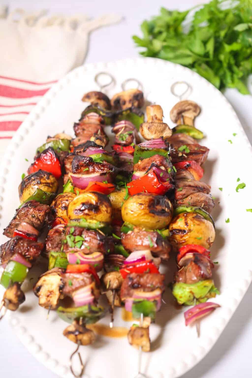 Grilled vegetable and meat skewers, including tender steak kabobs with mushrooms, potatoes, peppers, onions, and tomatoes, are served on a white plate and garnished with chopped herbs. Parsley and a napkin are in the background.