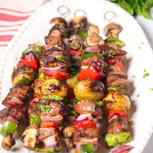 Grilled vegetable and meat skewers, including tender steak kabobs with mushrooms, potatoes, peppers, onions, and tomatoes, are served on a white plate and garnished with chopped herbs. Parsley and a napkin are in the background.