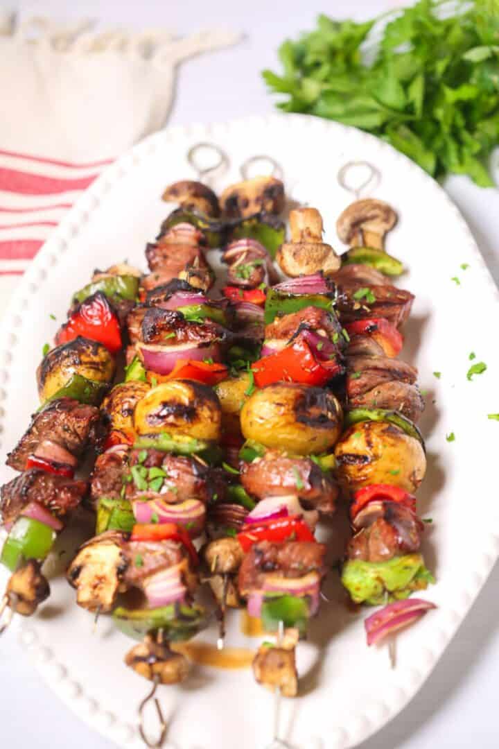 Grilled vegetable and meat skewers, including tender steak kabobs with mushrooms, potatoes, peppers, onions, and tomatoes, are served on a white plate and garnished with chopped herbs. Parsley and a napkin are in the background.