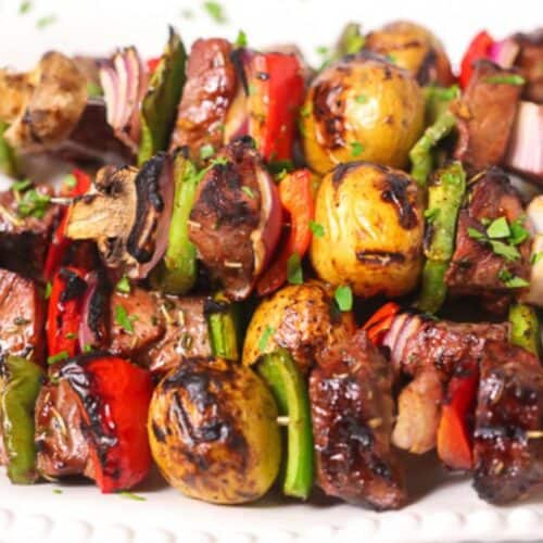Steak Kabobs with grilled beef, mushrooms, baby potatoes, bell peppers, and red onions are artfully arranged on a white plate and garnished with chopped herbs.