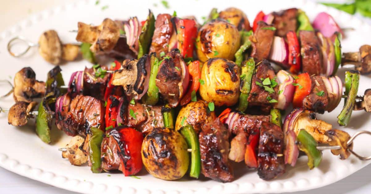 Steak Kabobs with grilled beef, mushrooms, baby potatoes, bell peppers, and red onions are artfully arranged on a white plate and garnished with chopped herbs.
