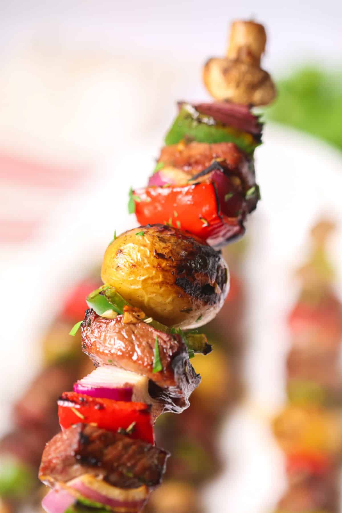 A close-up of steak kabobs showcases a grilled vegetable and meat skewer featuring mushrooms, peppers, onions, and herbs against a blurred background.