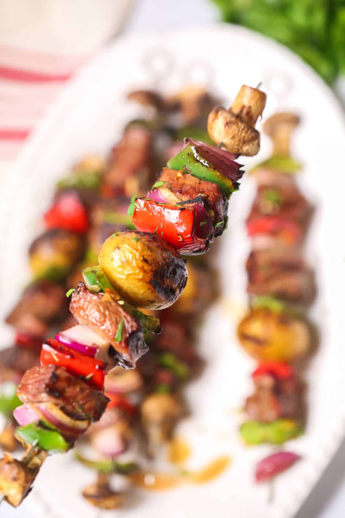 A Steak Kabob skewer with grilled mushrooms, bell peppers, onions, and tender meat is held above a platter with more skewers in the background.