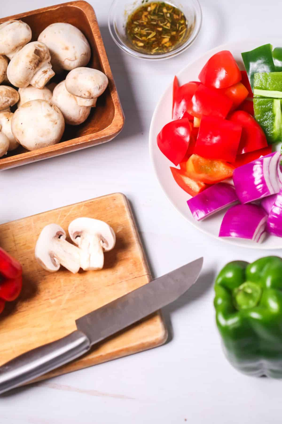 Chopped mushrooms, bell peppers, and red onion on a plate and cutting board with a knife, ready to be skewered for delicious Steak Kabobs; whole mushrooms in a bowl, and a small bowl of marinade sit nearby on a white surface.