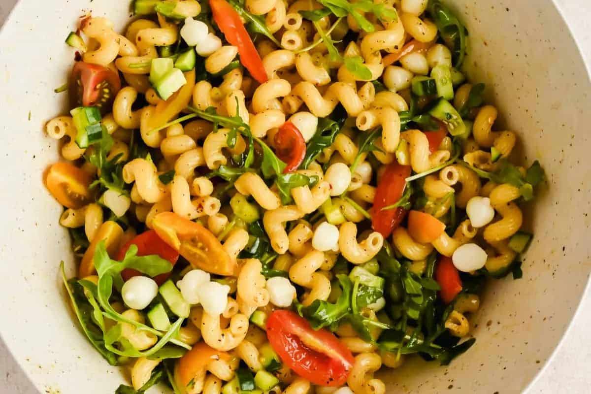 A bowl of pasta salad with cavatappi noodles, cherry tomatoes, arugula, diced cucumber, cheese pearls, and herbs—perfect for those who crave mouthwatering salads and refreshing summer salads.