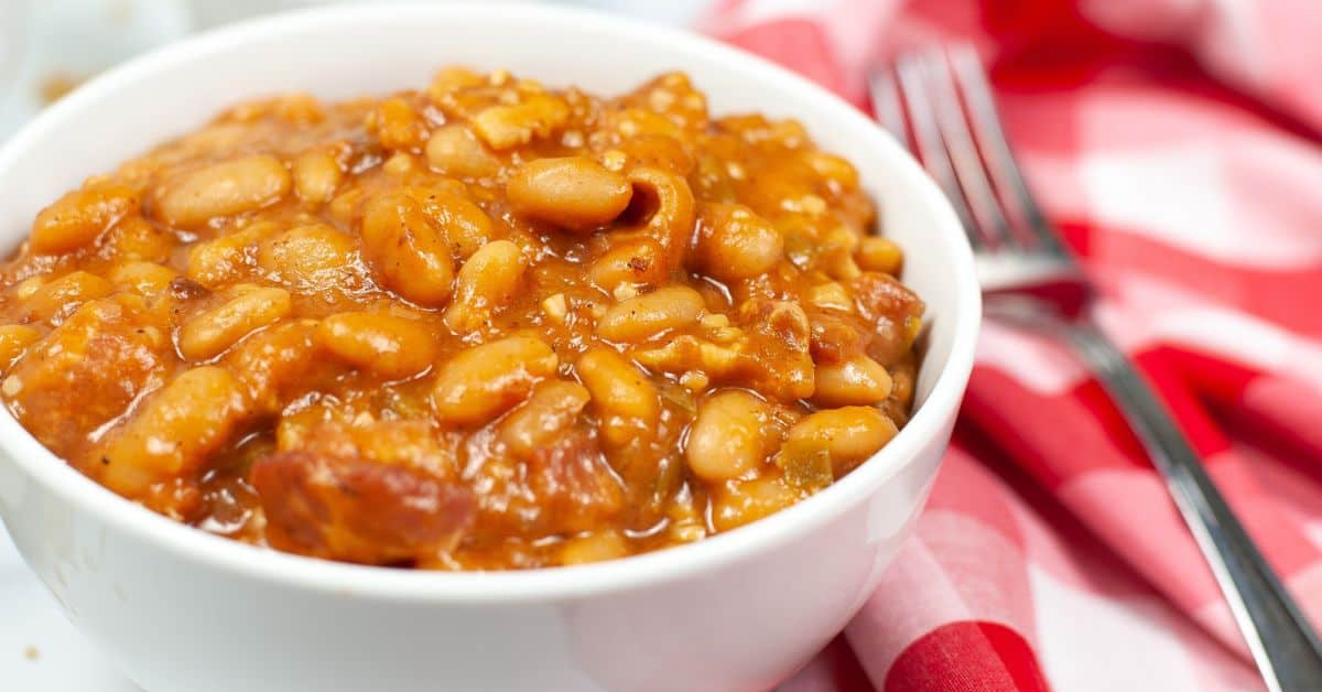 A white bowl filled with Instant Pot Baked Beans in sauce sits on a table next to a red and white checkered napkin and a fork.