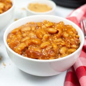 A white bowl filled with Instant Pot Baked Beans in a thick sauce, with a red and white checkered napkin nearby.