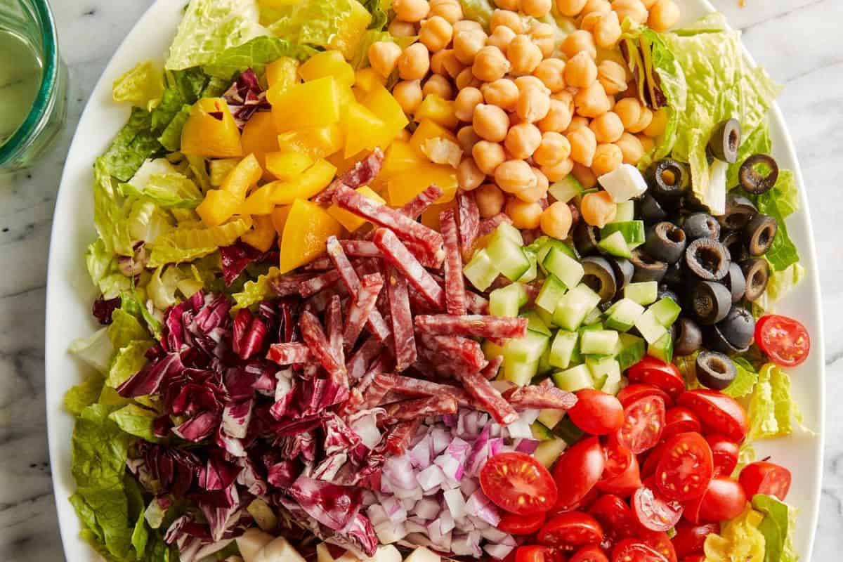 A mouthwatering salad with chopped lettuce, yellow bell pepper, chickpeas, salami strips, diced cucumbers, black olives, grape tomatoes, red onion, and radicchio on a white plate—perfect for fans of seasonal salads.