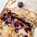 A Lemon Blueberry Loaf sits on a white plate, its glazed top dotted with fresh blueberries. Two slices are cut to reveal juicy berries inside, adding a zesty twist to this classic treat.
