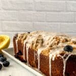 A Lemon Blueberry Loaf sits on a white platter, its glazed surface glistening. Garnished with blueberries and a halved lemon nearby, this rectangular bread rests elegantly on a white surface.