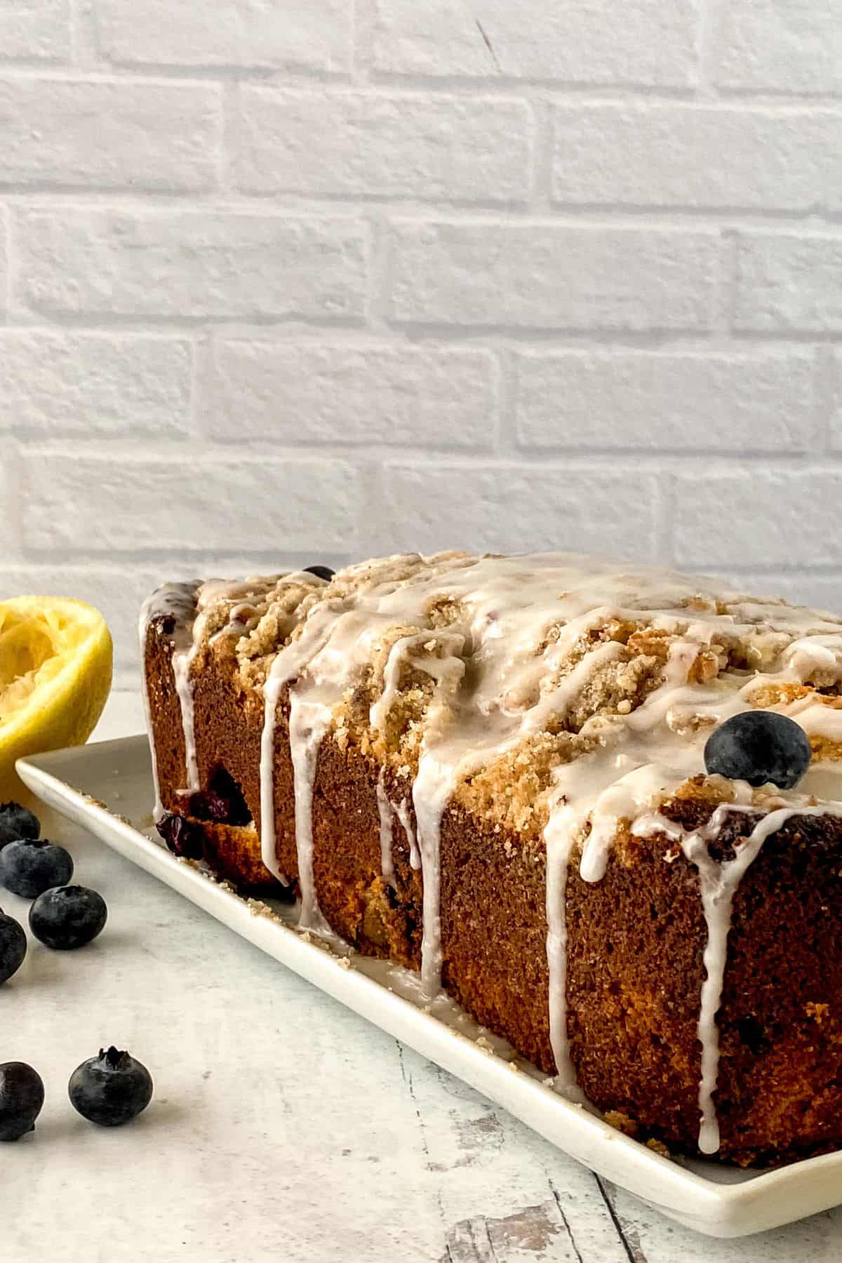 A Lemon Blueberry Loaf sits on a white platter, its glazed surface glistening. Garnished with blueberries and a halved lemon nearby, this rectangular bread rests elegantly on a white surface.