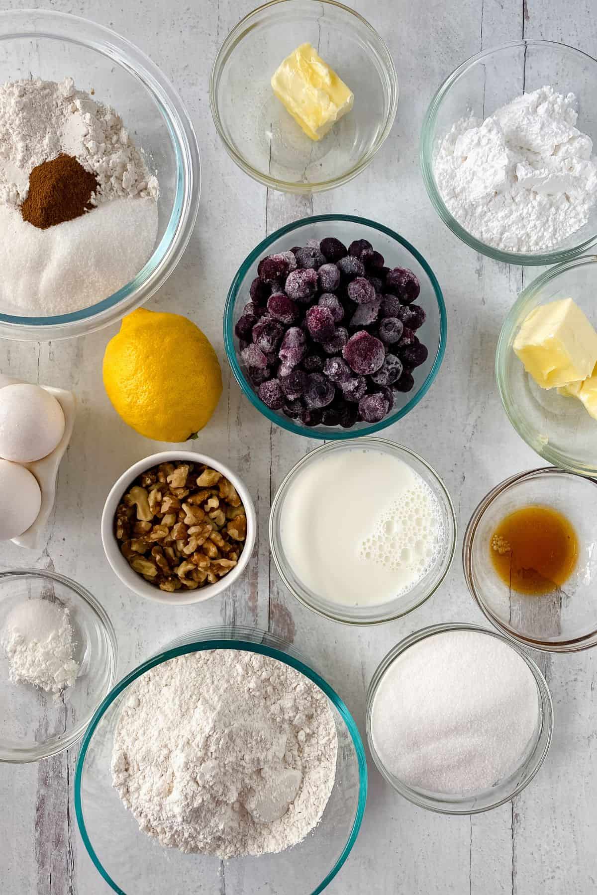 Assorted baking ingredients in glass bowls, including flour, sugar, eggs, butter, milk, vanilla, walnuts, lemon for a Lemon Blueberry Loaf, frozen cherries, and spices, arranged on a white surface.