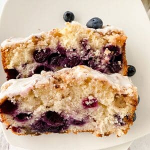 Two slices of Lemon Blueberry Loaf with white icing are stacked on a white plate, with a few whole blueberries beside them.