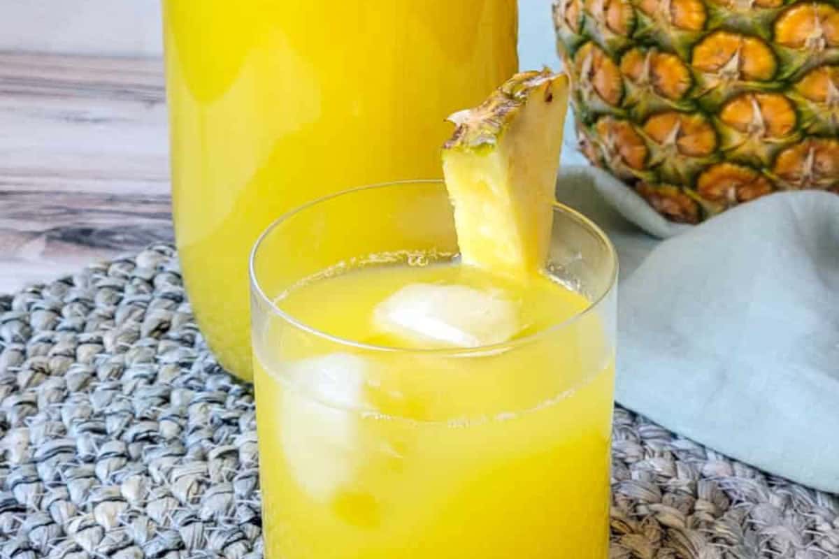 A glass of mouthwatering yellow pineapple juice with ice and a pineapple wedge sits on a woven mat; a glass jar of juice and a whole pineapple complete this must-try scene for fans of delicious pineapple recipes.