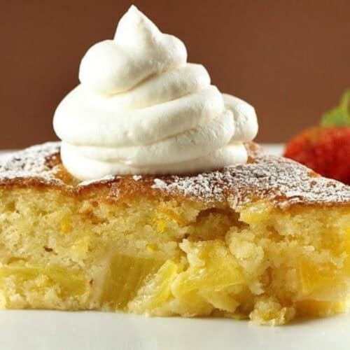 A slice of pineapple cake topped with a swirl of whipped cream and sprinkled with powdered sugar, with a strawberry in the background—a must try for fans of mouthwatering recipes.