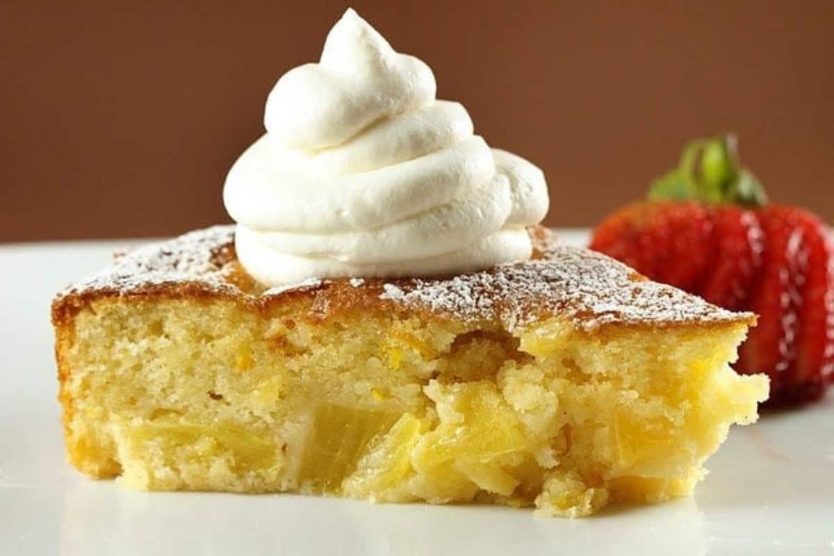 A slice of pineapple cake topped with a swirl of whipped cream and sprinkled with powdered sugar, with a strawberry in the background—a must try for fans of mouthwatering recipes.