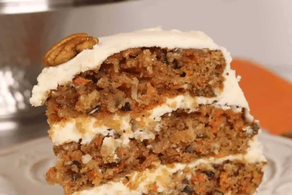 A close-up of a slice of carrot cake with cream cheese frosting, topped with a pecan and displayed on a white plate—a must try for fans of mouthwatering recipes.