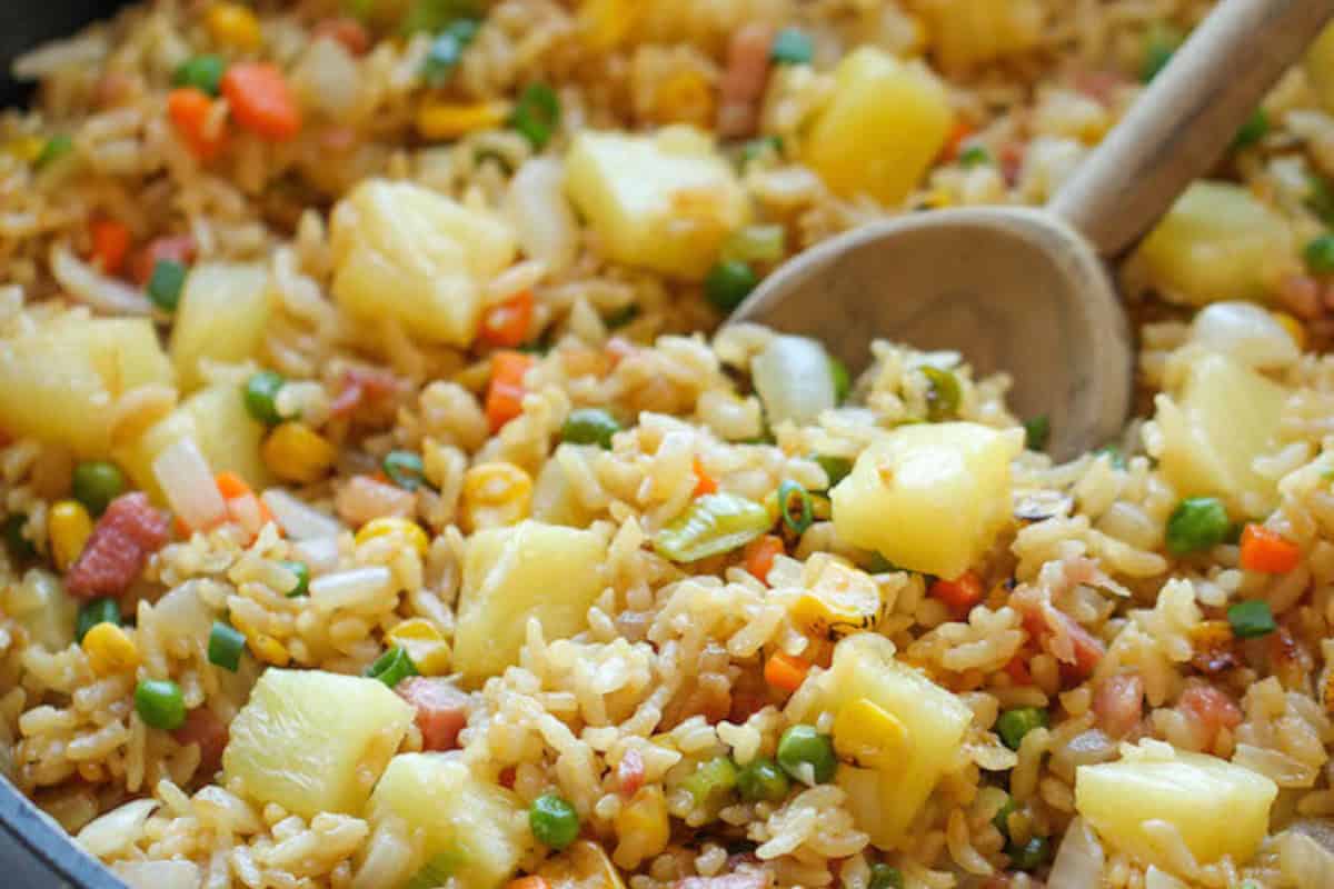 A close-up of fried rice mixed with diced pineapple, peas, carrots, corn, onions, and small pieces of ham, with a wooden spoon in the dish—one of the most mouthwatering pineapple recipes to try.
