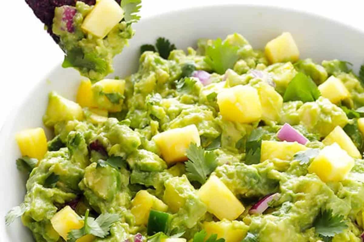A bowl of guacamole with chunks of avocado, diced mango, red onion, cilantro, and a chip dipping into it—one of those mouthwatering recipes you won’t want to miss.