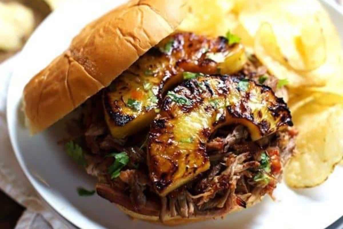 A must try pulled pork sandwich topped with grilled pineapple slices and fresh herbs, served with potato chips on a white plate—perfect for fans of mouthwatering recipes and creative pineapple recipes.