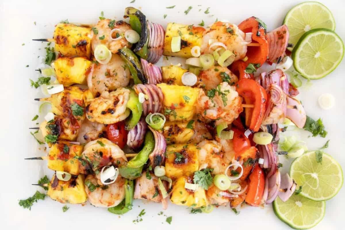 Grilled shrimp and vegetable skewers with pineapple, red onion, bell peppers, and herbs—one of the must try mouthwatering recipes—garnished with lime slices and chopped scallions on a white plate.
