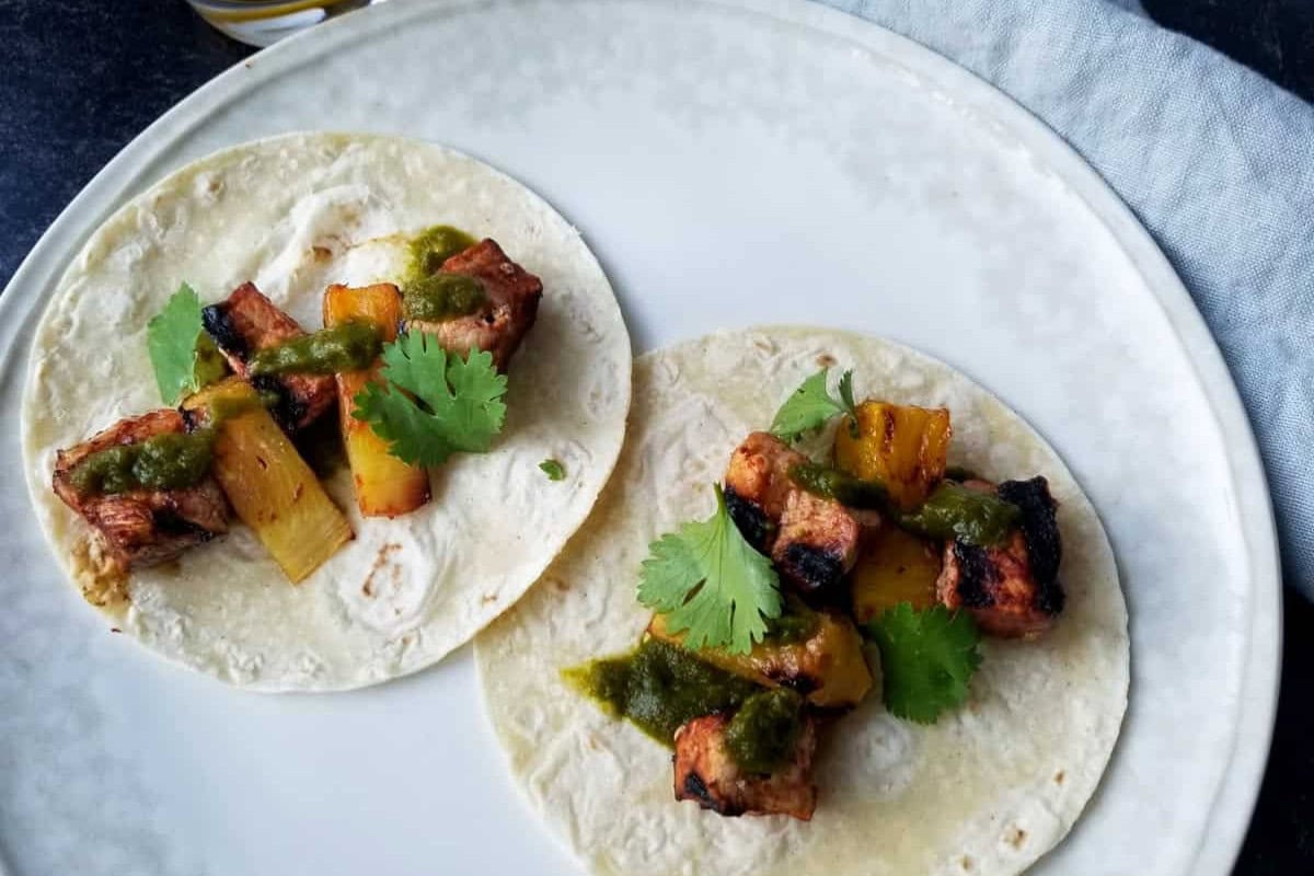 Two tacos on a white plate, each filled with grilled pork, pineapple chunks, green sauce, and cilantro on flour tortillas—a must try recipe for fans of mouthwatering recipes.