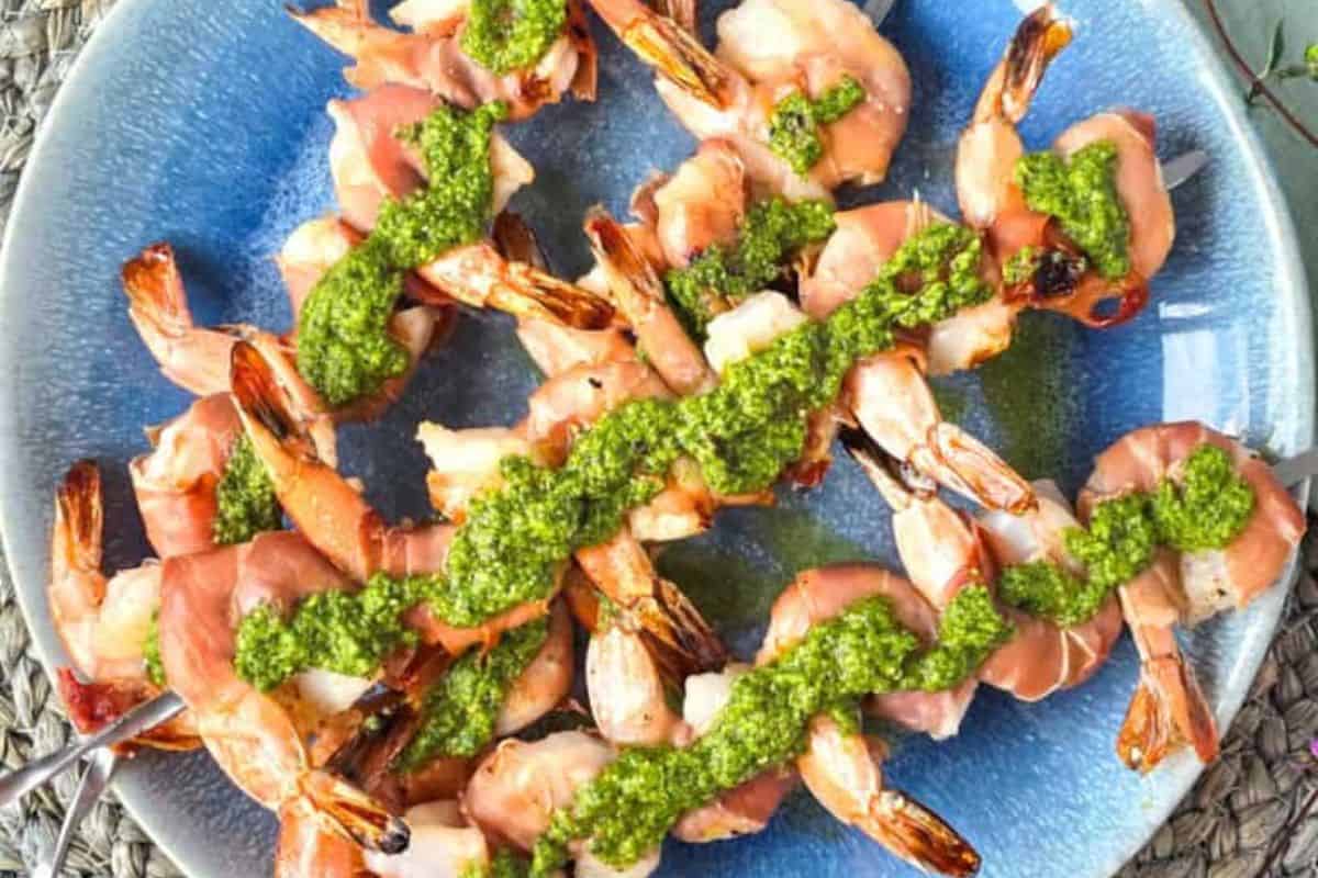 Grilled shrimp kabob skewers wrapped in prosciutto, topped with green pesto sauce, served on a blue plate—perfect for fans of kabob recipes.