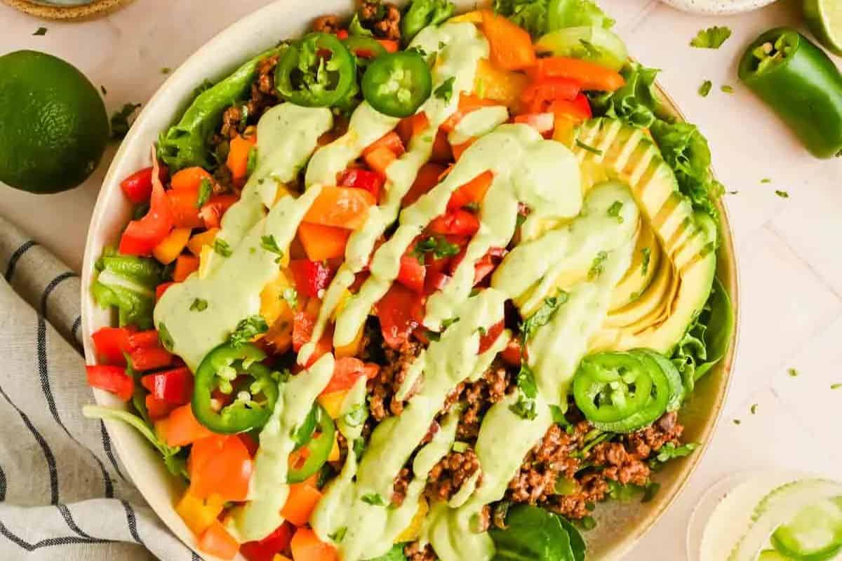 A bowl of taco salad with ground beef, sliced bell peppers, avocado, jalapeño, and green dressing on a bed of lettuce—perfect for those who crave mouthwatering salads.