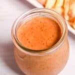 A glass jar filled with creamy, orange-colored Red Robin Campfire Sauce sits on a white surface, with a plate of French fries in the background.