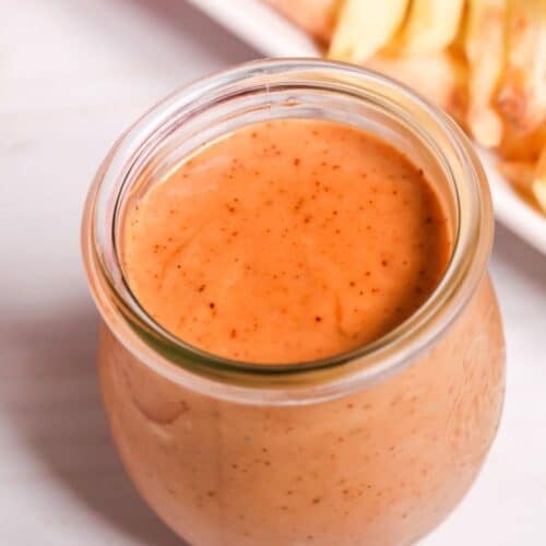A glass jar filled with creamy, orange-colored Red Robin Campfire Sauce sits on a white surface, with a plate of French fries in the background.