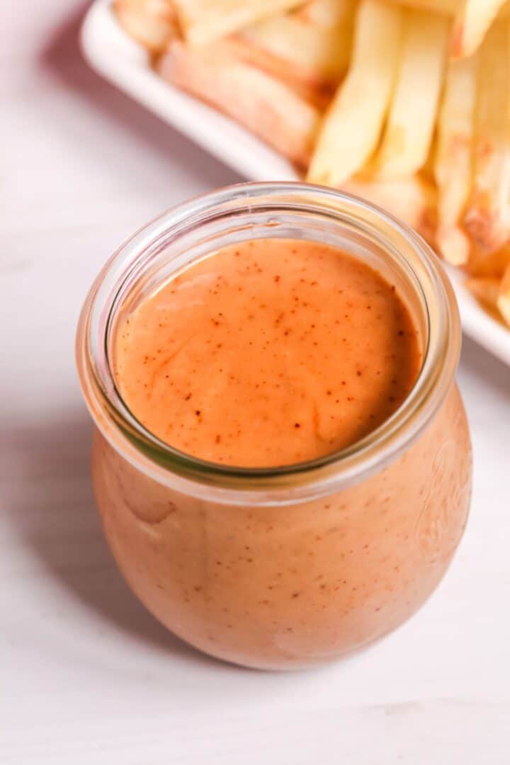 A glass jar filled with creamy, orange-colored Red Robin Campfire Sauce sits on a white surface, with a plate of French fries in the background.