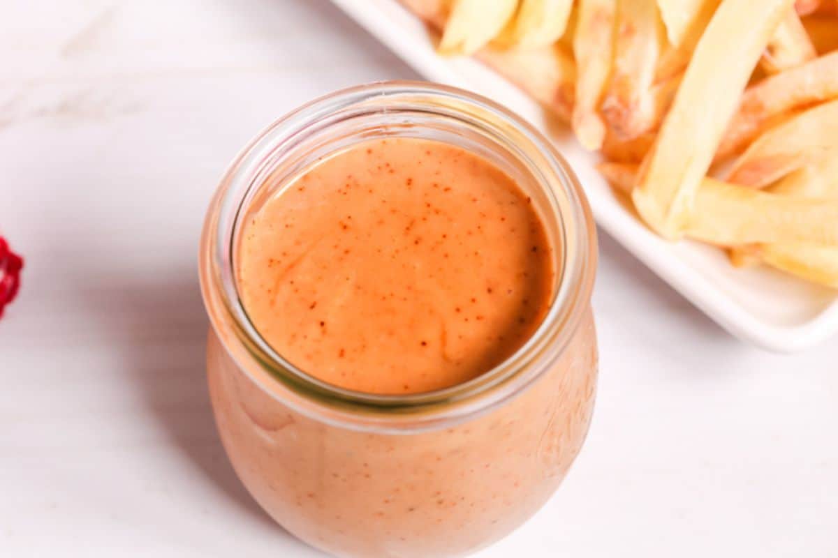 A small glass jar filled with light orange Red Robin Campfire Sauce sits next to a plate of French fries on a white surface.