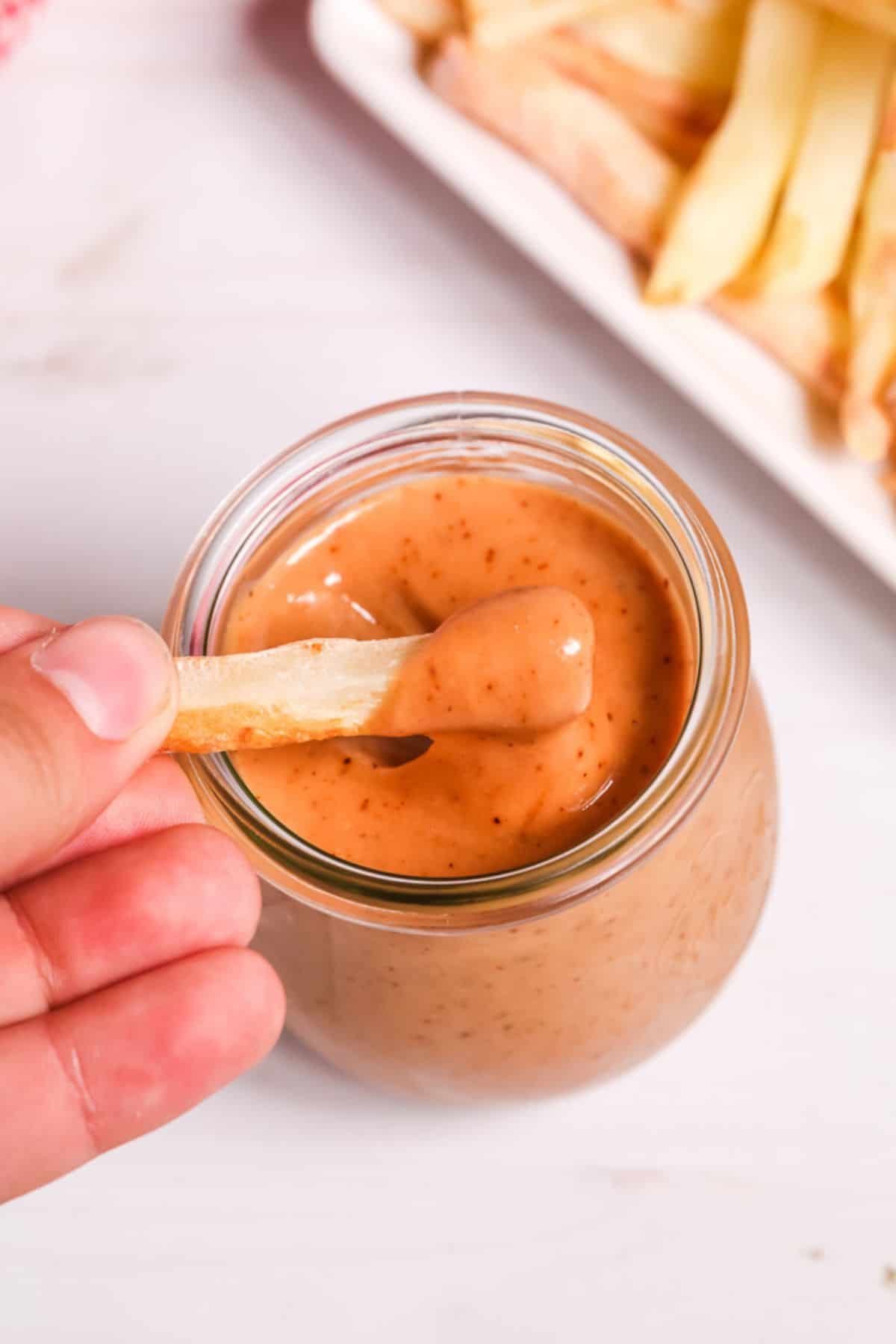 A hand dips a French fry into a jar. A plate of fries is visible in the background.