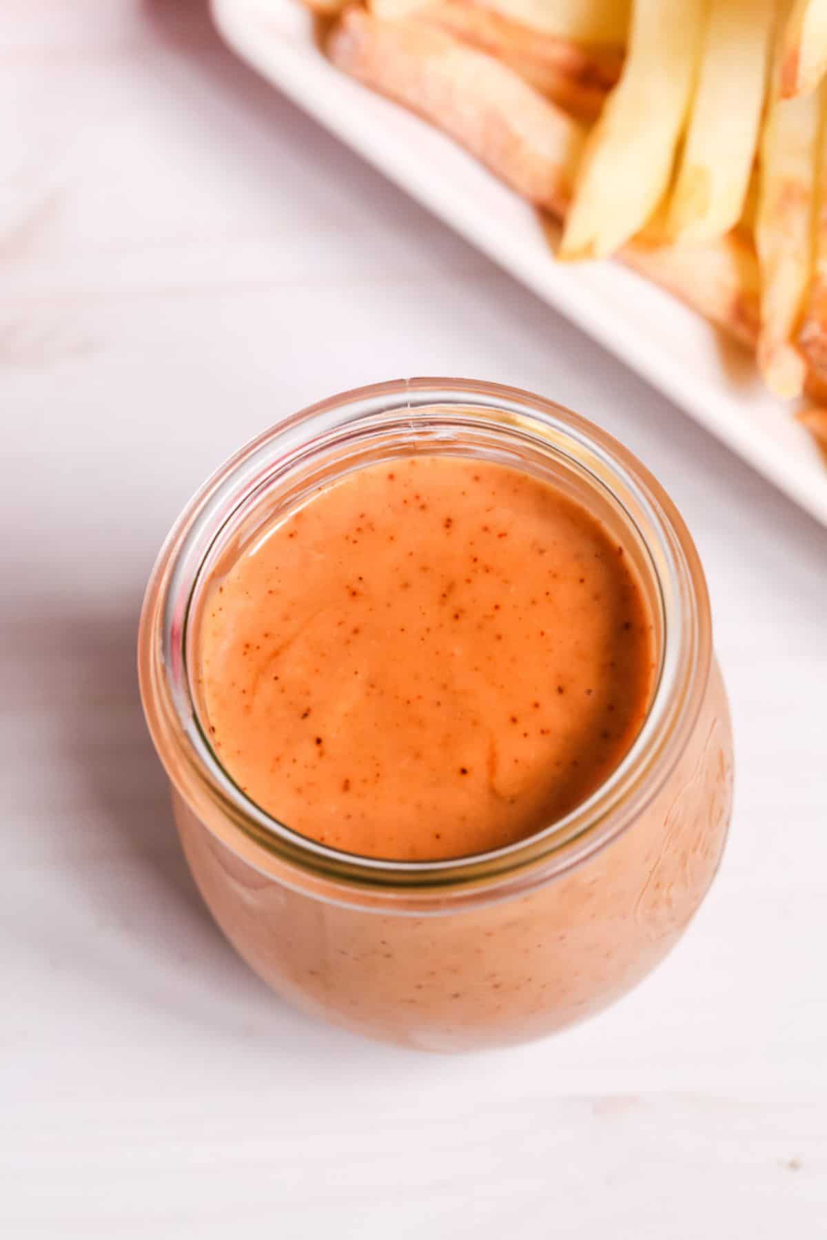 A glass jar filled with a creamy, orange dipping sauce—reminiscent of Red Robin Campfire Sauce—sits on a white surface, with a plate of French fries in the background.