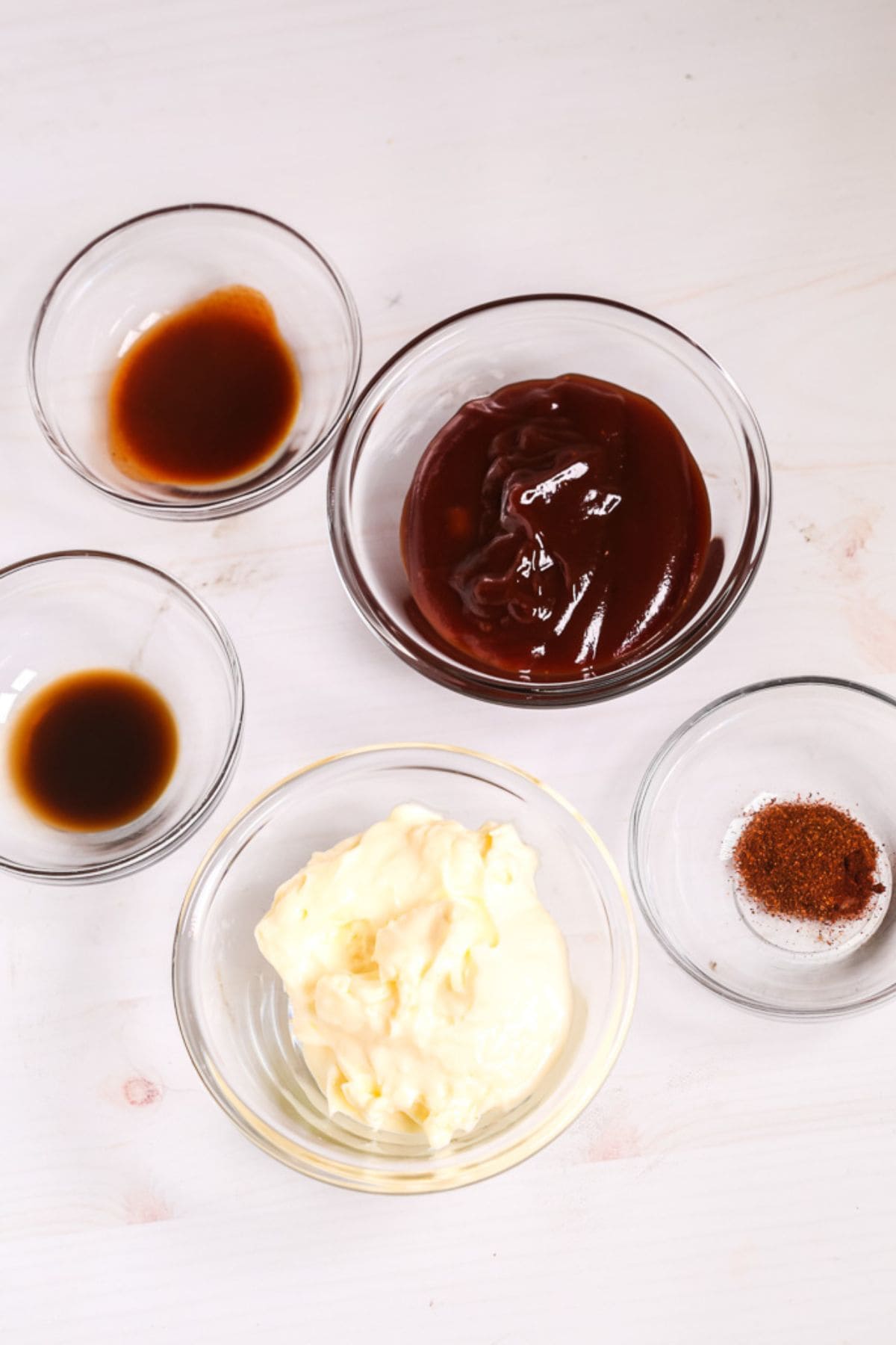 Five glass bowls on a white surface containing barbecue sauce, mayonnaise, Red Robin Campfire Sauce, a dark liquid, and a small amount of reddish-brown spice.