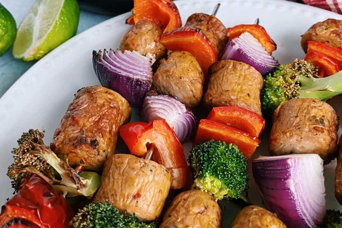 Vegetable and sausage kabob skewers with broccoli, red onion, red bell pepper, and sausage pieces arranged on a white plate. Lime wedges are visible in the background—perfect for those seeking flavorful kabob recipes.