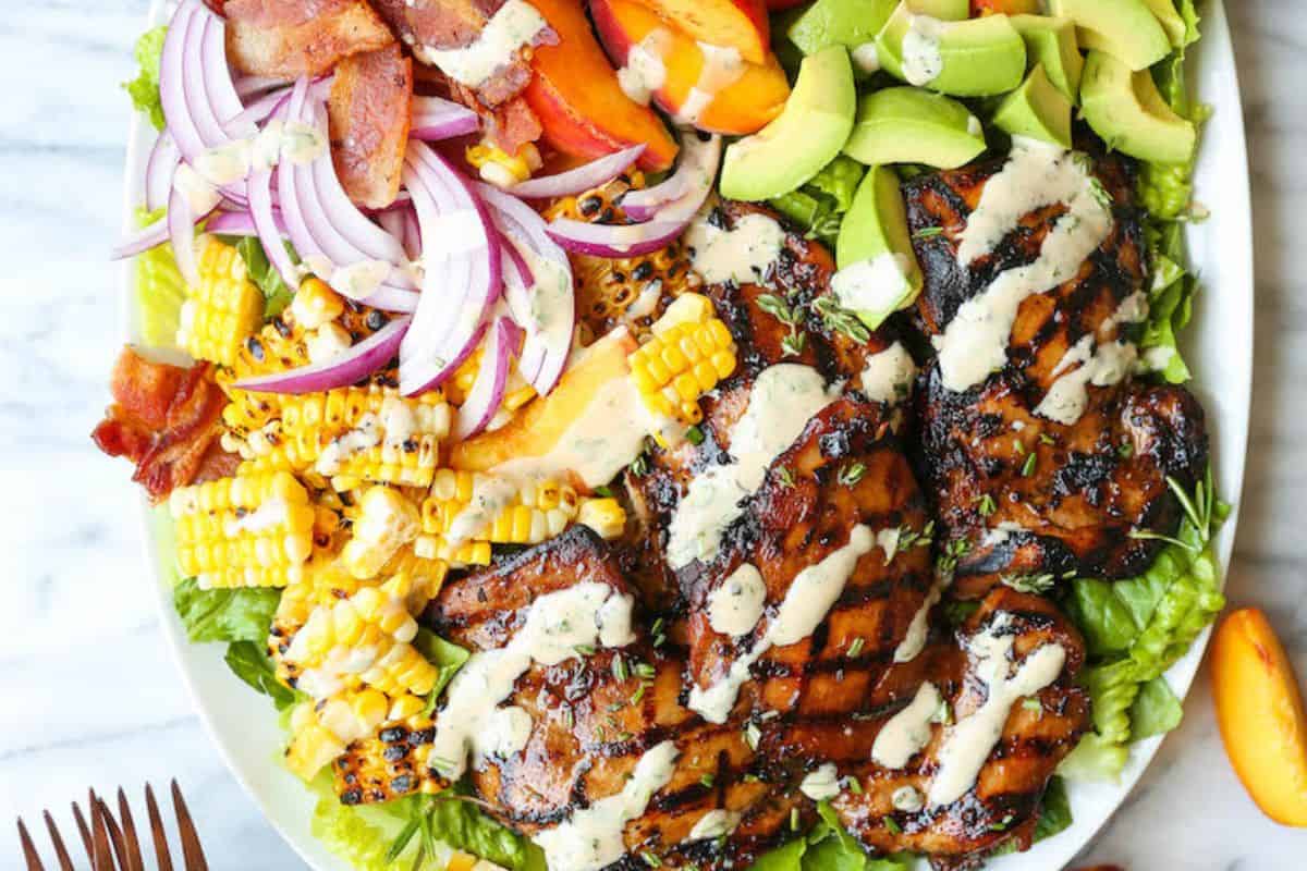 A plate of grilled chicken with creamy dressing, avocado, grilled corn, red onion, bacon, tomato, and lettuce arranged on a white dish—perfect for those who crave mouthwatering salads.