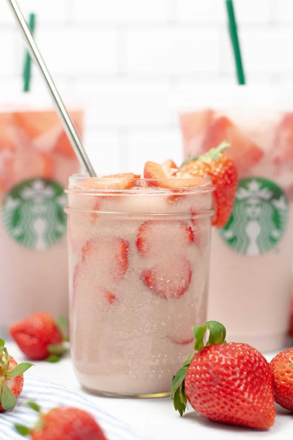 A glass jar filled with iced pink drink and sliced strawberries sits in front of two Starbucks cups featuring the signature Starbucks Pink Drink, with fresh strawberries nearby.
