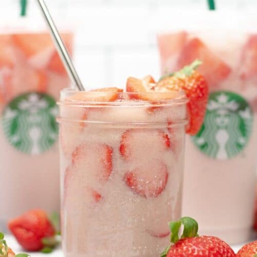A glass jar filled with iced pink drink and sliced strawberries sits in front of two Starbucks cups featuring the signature Starbucks Pink Drink, with fresh strawberries nearby.