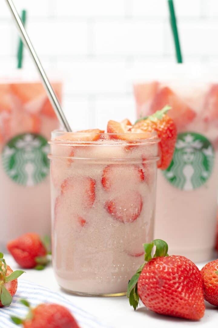A glass jar filled with iced pink drink and sliced strawberries sits in front of two Starbucks cups featuring the signature Starbucks Pink Drink, with fresh strawberries nearby.
