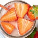 A glass filled with ice water, garnished with sliced strawberries and a whole strawberry, with a metal straw—this refreshing creation evokes the vibe of a Starbucks Pink Drink.