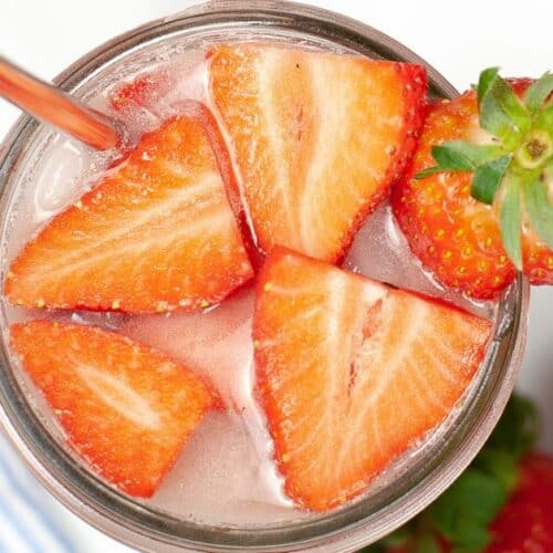 A glass filled with ice water, garnished with sliced strawberries and a whole strawberry, with a metal straw—this refreshing creation evokes the vibe of a Starbucks Pink Drink.