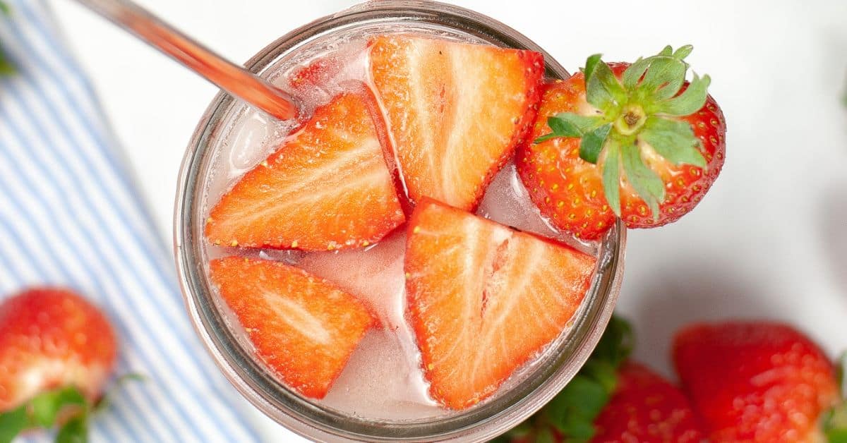 A glass filled with ice water, garnished with sliced strawberries and a whole strawberry, with a metal straw—this refreshing creation evokes the vibe of a Starbucks Pink Drink.