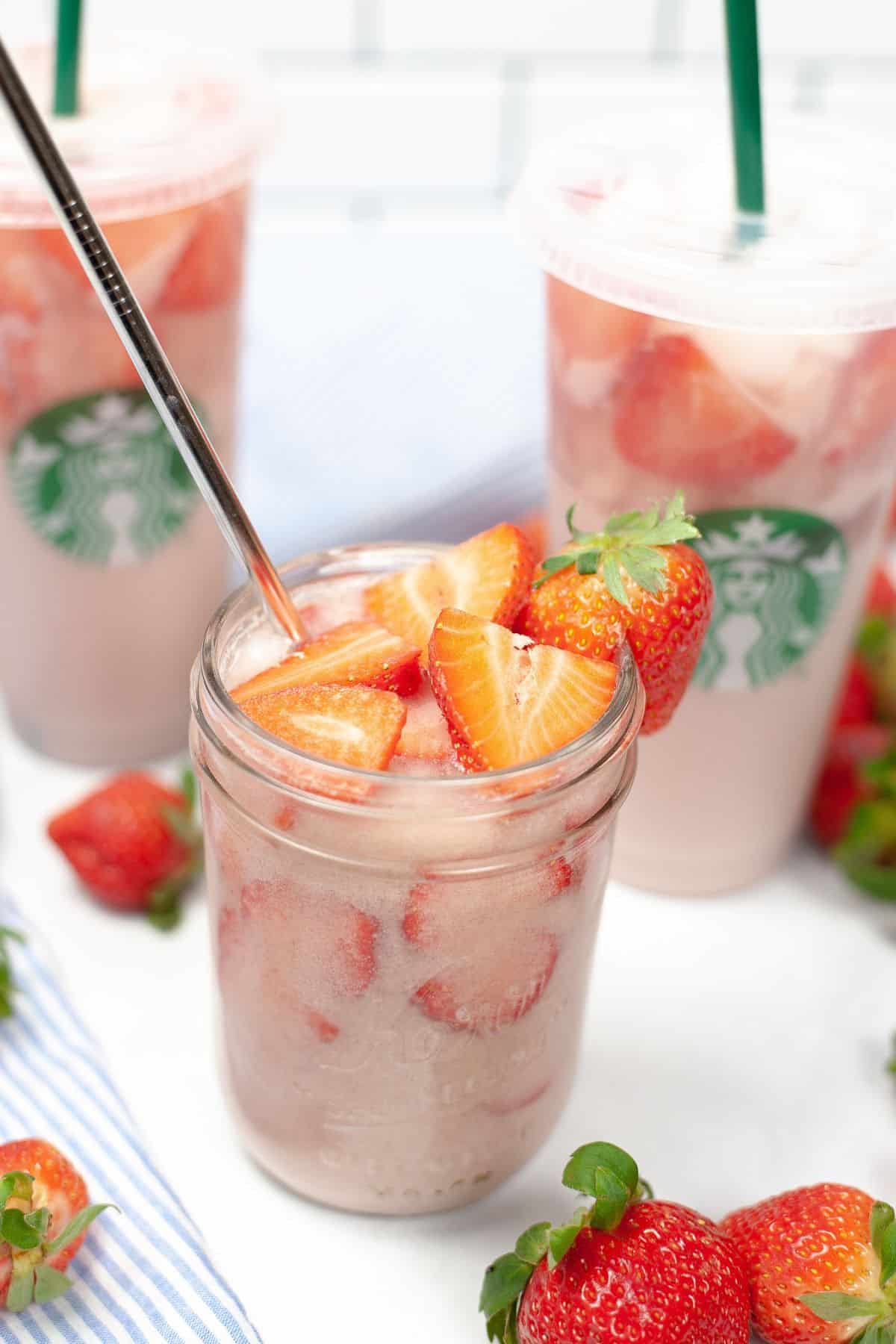 A mason jar and two Starbucks cups filled with the iconic Pink Drink and sliced strawberries, surrounded by fresh strawberries on a white surface.