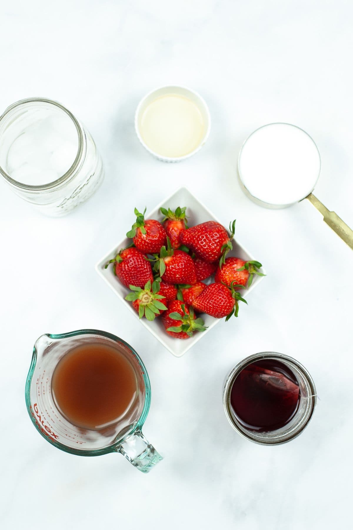 Top view of fresh strawberries in a bowl surrounded by a glass of juice, a glass of red liquid reminiscent of the popular Starbucks Pink Drink, a jar, a cup of milk, and a small bowl of liquid on a white surface.