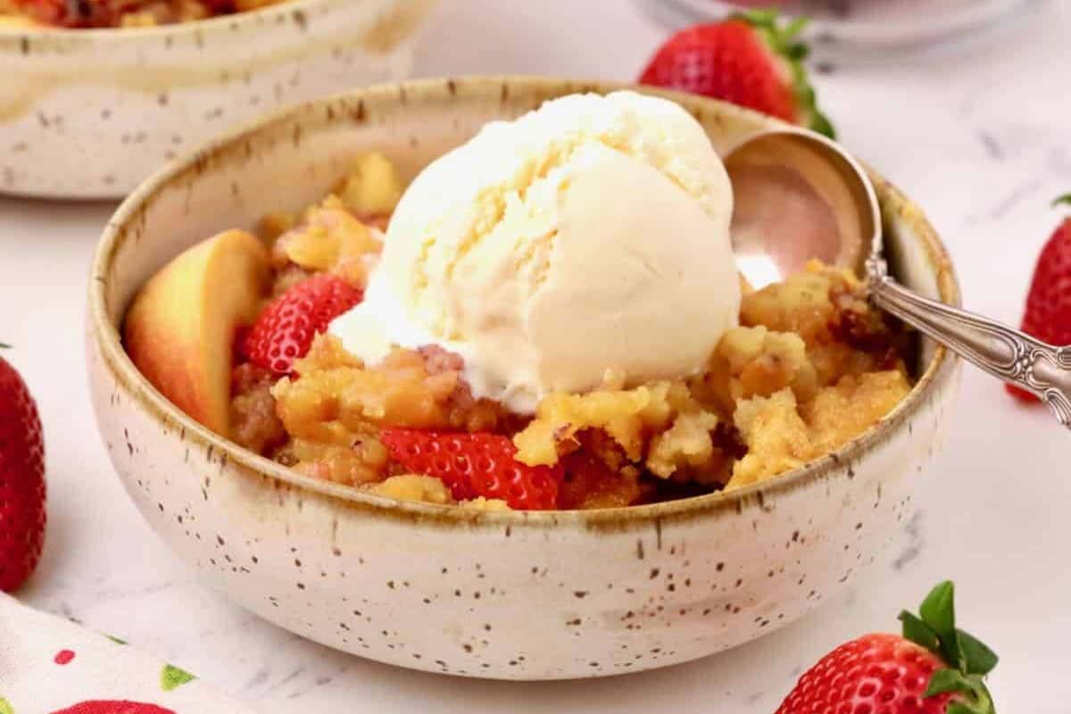 Close-up of a bowl of strawberry peach cobbler, garnished with peaches and strawberries. Topped with a scoop of vanilla ice cream.