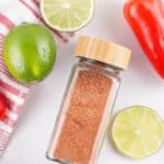 Glass jar with a wooden lid containing Chili Lime Seasoning surrounded by lime slices, a whole lime, a red bell pepper, a red kitchen towel, and measuring spoons.