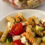 A bowl of rotini pasta salad featuring tri-color rotini tossed with cherry tomatoes, cucumber, red onion, and cubes of feta cheese. In the background, a larger serving bowl filled with the same vibrant rotini pasta salad completes the inviting scene.